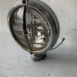 May include: A vintage chrome car headlight with a clear glass lens and a wire attached. The headlight is on a white textured surface.
