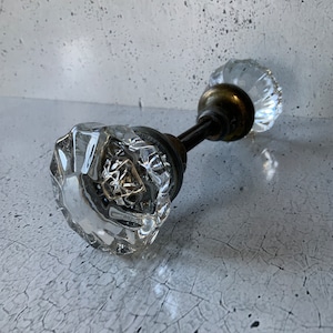Może przedstawiać: Przezroczysta szklana gałka do drzwi o fasetowym wzorze. Gałka ma podstawę i trzpień w kolorze mosiądzu. Szkło jest przezroczyste i odbija światło. Gałka do drzwi jest prawdopodobnie zabytkowa lub antyczna.