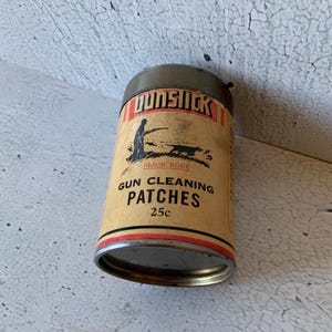 Puede incluir: Una lata vintage de parches de limpieza para armas Gun Slick. La lata es amarilla con detalles rojos y negros. Presenta una ilustración en blanco y negro de un cazador con un perro. El texto en la lata dice "Gun Slick", "Slick Bore", "Gun Cleaning Patches" y "25c".