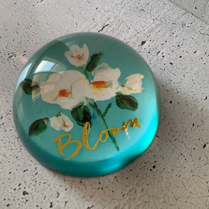 May include: A teal blue glass paperweight with a white floral design and the word "Bloom" in gold lettering.