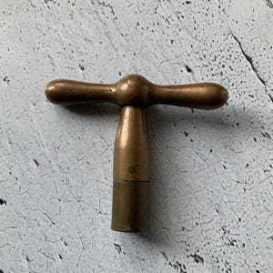 Pode incluir: Um tampão de garrafa de cor bronze com uma pega em forma de T. A pega tem extremidades arredondadas e o tampão tem uma base cilíndrica. O metal tem um aspeto ligeiramente envelhecido, sugerindo que é vintage ou antigo.