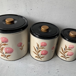 Peut inclure: Ensemble de trois bocaux de cuisine vintage. Les bocaux sont blanc cassé avec un couvercle noir et un bouton doré. Chaque bocal présente un motif floral rose avec des tiges et des feuilles marron. Les bocaux sont de tailles différentes.