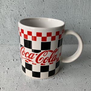 May include: White ceramic mug with a black and red checkered pattern and the Coca-Cola logo in red.