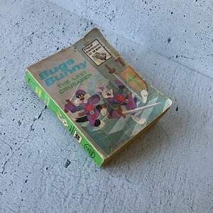 Puede incluir: Un cómic vintage de Bugs Bunny titulado "El Último Cruzado" con una portada verde que presenta a Bugs Bunny y un gato negro con trajes de caballeros medievales. La portada del cómic tiene un pequeño anuncio de "Flip-a-Cartoons See 'em Move!"