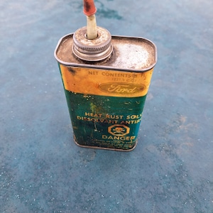 May include: A vintage green and yellow metal can with a red and white spout. The can is labelled "Ford Heat Rust Solvent Dissolvant Antirouille" and has a skull and crossbones with the word "Danger" printed below it.