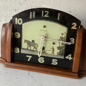 Könnte beinhalten: Vintage Holz-Kaminuhr mit schwarzem Zifferblatt und hellgrünem Hintergrund. Die Uhr zeigt eine Silhouette von Pferd und Reiter, mit dem Wort "BERTMAR" sichtbar. Die Uhr hat einen braunen Holzrahmen und weiße Ziffern.