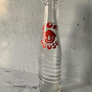 May include: A clear glass soda bottle with a red and white label that says "Pop House" with a cartoon house with a smiling face. The bottle has a textured surface.