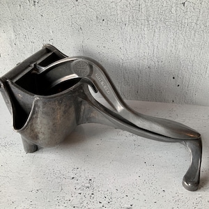 Puede incluir: Exprimidor de cítricos plateado de época. El exprimidor tiene un brazo de palanca y un receptáculo en forma de cuenco para recoger el zumo. Las palabras "MADE IN U.S.A." son visibles en el mango. El exprimidor es de metal y tiene un aspecto antiguo y desgastado.