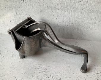 May include: A vintage, silver-toned citrus juicer. The juicer has a lever arm and a bowl-shaped receptacle for catching juice. The words "MADE IN U.S.A." are visible on the handle. The juicer is made of metal and has a worn, antique appearance.