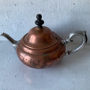 May include: A copper teapot with a silver handle and a wooden knob on the lid. The teapot has a rounded body and a slightly flared spout.