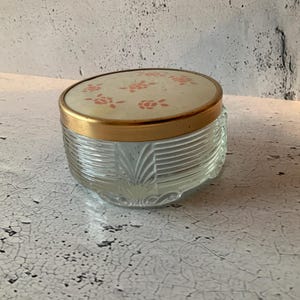 May include: A clear glass jar with a gold lid and a floral design on the top. The jar has a textured pattern on the sides.