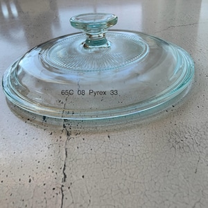 Op de afbeelding: Een doorzichtig glazen deksel met een knop aan de bovenkant. Het deksel heeft een verhoogde rand en de tekst "65C 08 Pyrex 33" gedrukt op de onderkant.