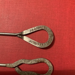Puede incluir: Dos ganchos para botas de metal antiguos. El gancho superior tiene el texto "A.D. SHUTE BOOT FACTOR WAKEFIELD" grabado. El gancho inferior tiene un patrón decorativo de espiral.