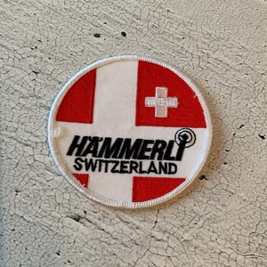 May include: Round patch with a white border, featuring the Swiss flag design in red and white. The text "HAMMERLI SWITZERLAND" is printed in black, with a logo of a target and a circle. The patch is likely for a sports or outdoor activity.