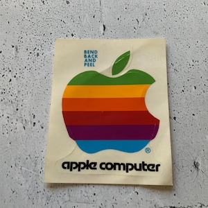 Peut inclure: Un autocollant rectangulaire présentant le logo Apple classique aux rayures arc-en-ciel avec une feuille verte. Au-dessus du logo, le texte indique BEND BACK AND PEEL. Sous le logo, le texte indique apple computer en police noire minuscule.