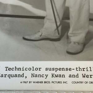 May include: Black and white photo of a person's legs wearing white pants and white shoes. The person is holding a cane. The photo is from a movie poster and includes the text "Bros.' Technicolor suspense-thriller, tian Marquand, Nancy Kwan and Werner 1967 BY WARNER BROS. PICTURES INC. COUNTRY OF ORIGIN U.S.A."