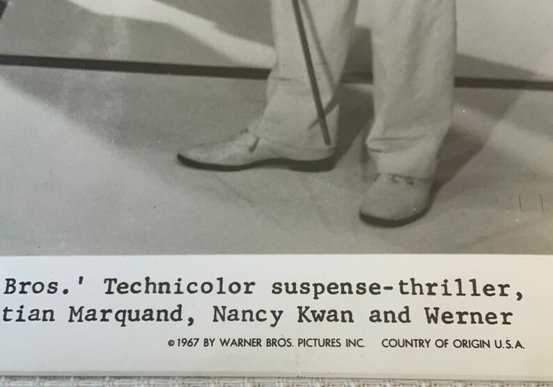 May include: Black and white photo of a person's legs wearing white pants and white shoes. The person is holding a cane. The photo is from a movie poster and includes the text "Bros.' Technicolor suspense-thriller, tian Marquand, Nancy Kwan and Werner 1967 BY WARNER BROS. PICTURES INC. COUNTRY OF ORIGIN U.S.A."