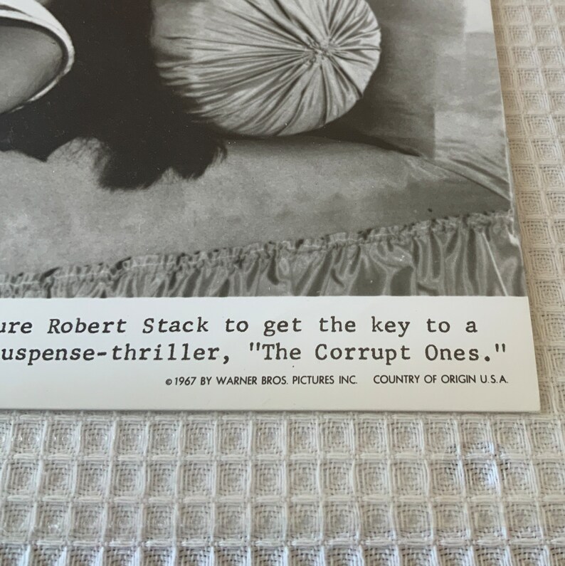 May include: Black and white photo of a woman lying on a bed with a pillow. The photo is from the movie "The Corrupt Ones" starring Robert Stack. The text on the photo reads: "ure Robert Stack to get the key to a suspense-thriller, "The Corrupt Ones." &copy; 1967 BY WARNER BROS. PICTURES INC. COUNTRY OF ORIGIN U.S.A."