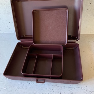 Puede incluir: Una caja de joyería de plástico color burdeos con tapa abatible y una bandeja extraíble con compartimentos. La caja tiene una pequeña manija en la parte delantera.