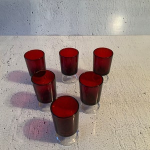 May include: Six small, clear glass shot glasses with a red, opaque base. The glasses are arranged in a circle on a white surface.