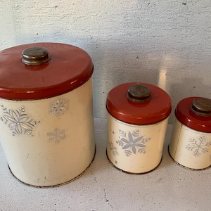 Puede incluir: Conjunto de tres botes de metal vintage con tapas rojas y diseños de copos de nieve blancos. Los botes son de diferentes tamaños y son perfectos para guardar alimentos básicos de la cocina.
