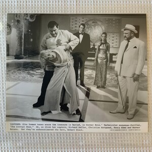 May include: A black and white photo from a movie scene. A man in a white suit is holding a cane. A woman in a long dress is being held by a man in a long robe. The text on the photo reads "TORTURED: Elke Sommer knows where the treasure is buried, in Warner Bros. Technicolor suspense-thriller, "The Corrupt Ones." Or, so think her captors, Richard Haller, Christian Marquand, Nancy Kwan and Werner Peters. But they've underestimated the hero, Robert Stack."