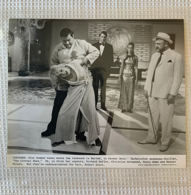 May include: A black and white photo from a movie scene. A man in a white suit is holding a cane. A woman in a long dress is being held by a man in a long robe. The text on the photo reads "TORTURED: Elke Sommer knows where the treasure is buried, in Warner Bros. Technicolor suspense-thriller, "The Corrupt Ones." Or, so think her captors, Richard Haller, Christian Marquand, Nancy Kwan and Werner Peters. But they've underestimated the hero, Robert Stack."