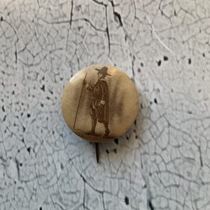 Könnte beinhalten: Ein Vintage-Knopf mit einer Schwarzweißillustration einer Person in Hut und Mantel, die einen Stab hält. Der Knopf ist rund und hat einen braunen Hintergrund.