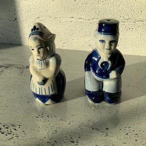 Może przedstawiać: Para niebiesko-białych ceramicznych solniczek i pieprzniczek w kształcie Holendrów w tradycyjnych strojach. Kobieta ma na sobie niebiesko-białą sukienkę i biały czepek. Mężczyzna ma na sobie niebieską kurtkę i spodnie oraz niebieski kapelusz.