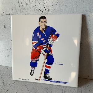 May include: A white ceramic tile featuring a vintage illustration of a hockey player in a blue and red uniform with the word "RANGERS" on the front. The player is holding a hockey stick and skating on the ice. The tile has the text "ANDY BATHGATE" and "H.M.COYMAN" printed on it.