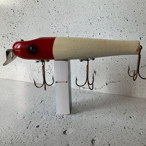 May include: A red and white wooden fishing lure with three treble hooks. The lure is approximately 25 cm long.