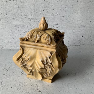 Op de afbeelding: Een decoratieve doos met een vierkante vorm en een versierd ontwerp. De doos is gemaakt van licht beige materiaal en heeft een bloemenpatroon. Het deksel van de doos heeft een klein, puntig knopje.