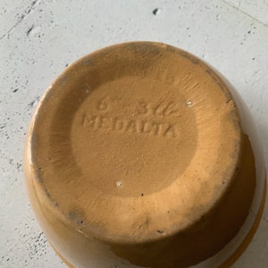 May include: A tan ceramic bowl with a brown glaze. The bottom of the bowl has the words "Made in USA" and "Meadow" stamped into the clay.