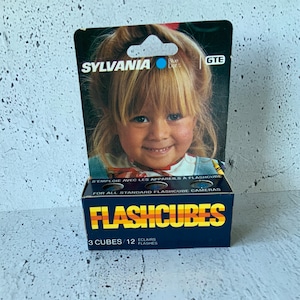 Peut inclure: Une boîte de flashcubes Sylvania Blue Dot bleue et noire. La boîte présente une photo d'une jeune fille souriante. Le texte sur la boîte indique "FLASHCUBES 3 CUBES/12 ECLAIRS FLASHES".