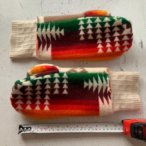 Può includere: Un paio di guanti in lana colorati con un motivo geometrico nei toni del verde, rosso, arancione e crema. I guanti hanno polsini a coste color crema. Un metro a nastro è posto accanto ai guanti. Misurano circa 26 cm.