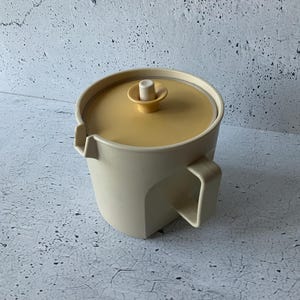 Puede incluir: Una jarra vintage de color crema con tapa y asa a juego. La tapa es de color amarillo claro con un pequeño asa cilíndrica. La jarra tiene un pico y un asa curvada.