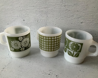 Puede incluir: Tres tazas de cerámica blanca con diseños florales y a cuadros verdes. Las tazas tienen un diseño clásico con un asa en el lateral. Las tazas están dispuestas en fila, mostrando sus diseños únicos.