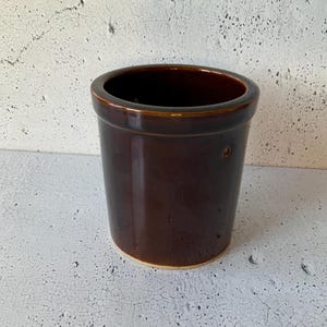 Puede incluir: Un tarro de cerámica marrón con una abertura ancha y un borde ligeramente acampanado. El tarro tiene un acabado liso y esmaltado y un brillo sutil.
