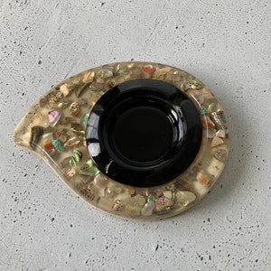 May include: A teardrop-shaped ashtray with a black centre and a clear outer layer filled with iridescent seashells.