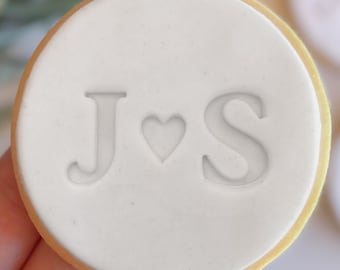 May include: White round cookie with the letters J and S and a heart symbol in between.