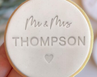 Könnte beinhalten: Runder weißer Keks mit grauer Glasur und dem Text "Mr & Mrs THOMPSON" und einem kleinen Herz.