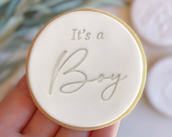 Może przedstawiać: Okrągłe białe ciasteczko z złotą obwódką i napisem "It's a Boy" w srebrnym kolorze.