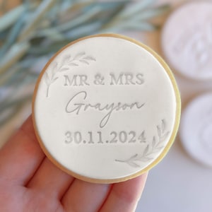 May include: White round cookie with a white icing design. The icing says "MR & MRS Grayson 30.11.2024" with a leafy design on either side.