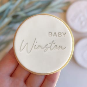 Könnte beinhalten: Ein runder weißer Keks mit goldfarbenem Rand, verziert mit dem Text "BABY Winston" in einer kursivschrift.