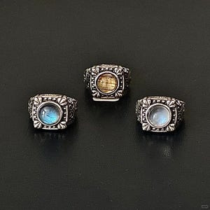 May include: Three ornate silver rings with square settings. Each ring features a different gemstone: a blue-green labradorite, a yellow citrine, and a white moonstone. The rings have detailed floral and scrollwork designs around the gemstones.