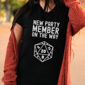 May include: A black t-shirt with a white graphic of a twenty-sided die and the text "New Party Member On The Way".