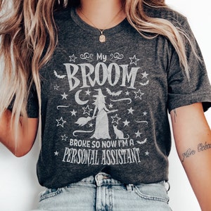 May include: Dark gray t-shirt with a witch graphic and the text "My Broom Broke So Now I'm a Personal Assistant." The design includes stars, bats, a cat, and a witch. A casual, Halloween-themed tee.