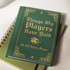May include: A green notebook with gold lettering that reads "Things My Players Have Said" and "So I'll Never Forget." A gold dragon illustration is centered below the title. The notebook is on top of a spiral-bound notebook.
