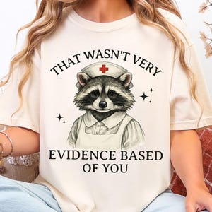 May include: Cream-colored t-shirt featuring a raccoon dressed as a nurse, wearing a nurse's cap with a red cross. The text above the raccoon reads "THAT WASN'T VERY" and below reads "EVIDENCE BASED OF YOU".