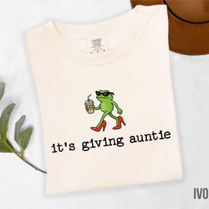May include: Ivory-colored t-shirt with the text "it's giving auntie." The shirt features a cartoon frog wearing sunglasses and red high heels, holding a coffee drink. The shirt is made by Comfort Colors.